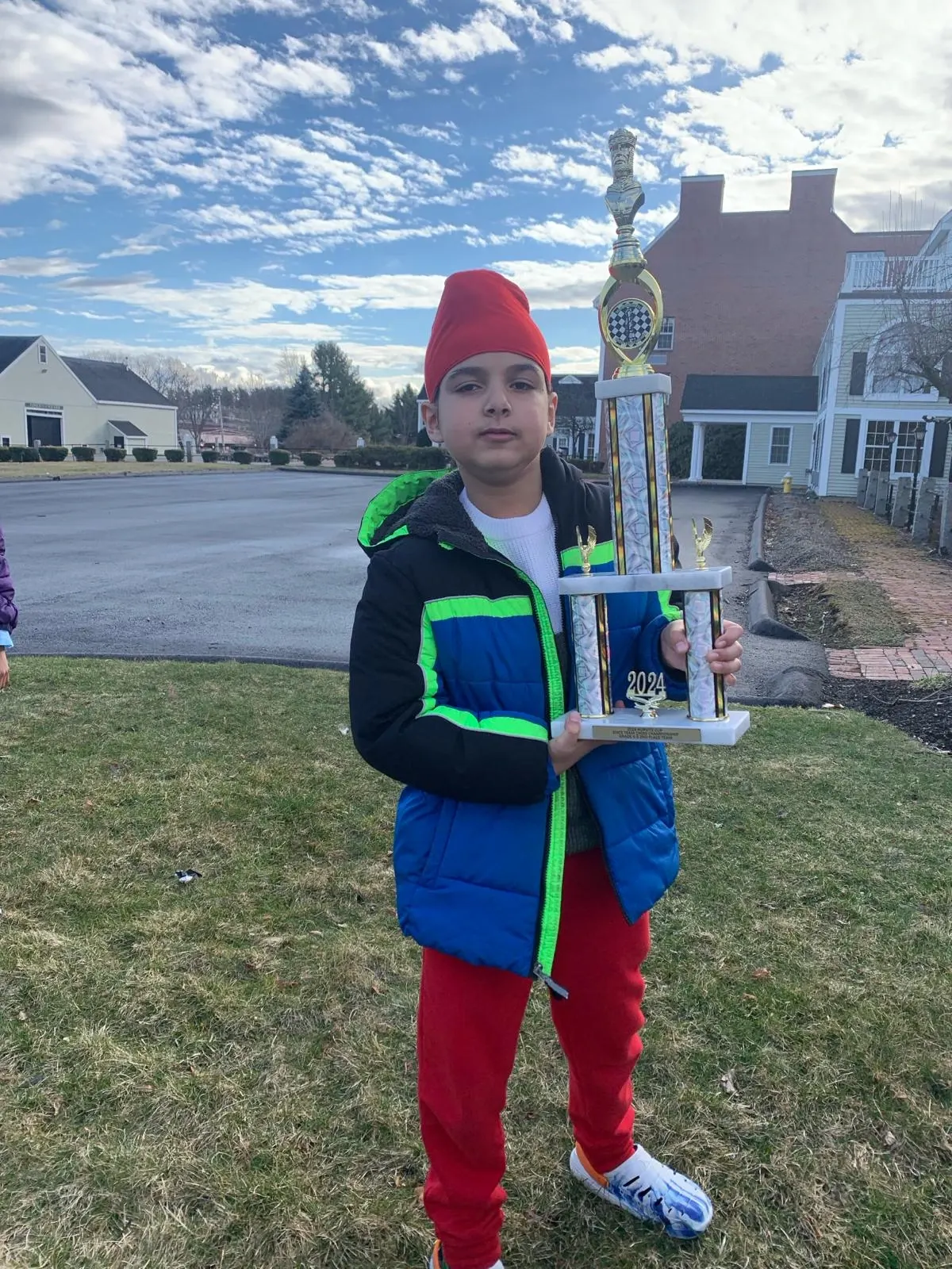 A young boy in a red turban and winter jacket proudly holding a tall,national chess trophy from outdoors to celebrate his tournament achievement.