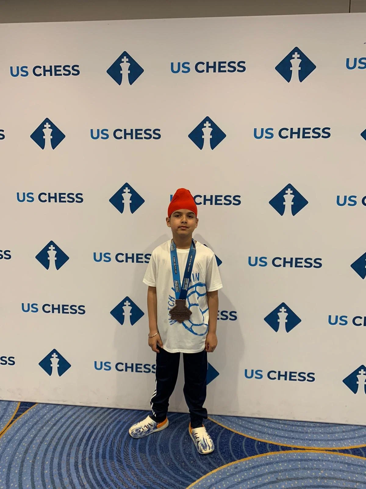 A young boy wearing a red turban holding large bronze chess medal  from national middle school champioship k8 standing in front of a US Chess step-and-repeat banner