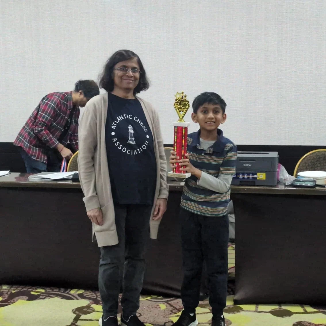 A young boy holding a tall red and gold chess trophy, posing alongside a representative wearing an Atlantic Chess Association shirt.