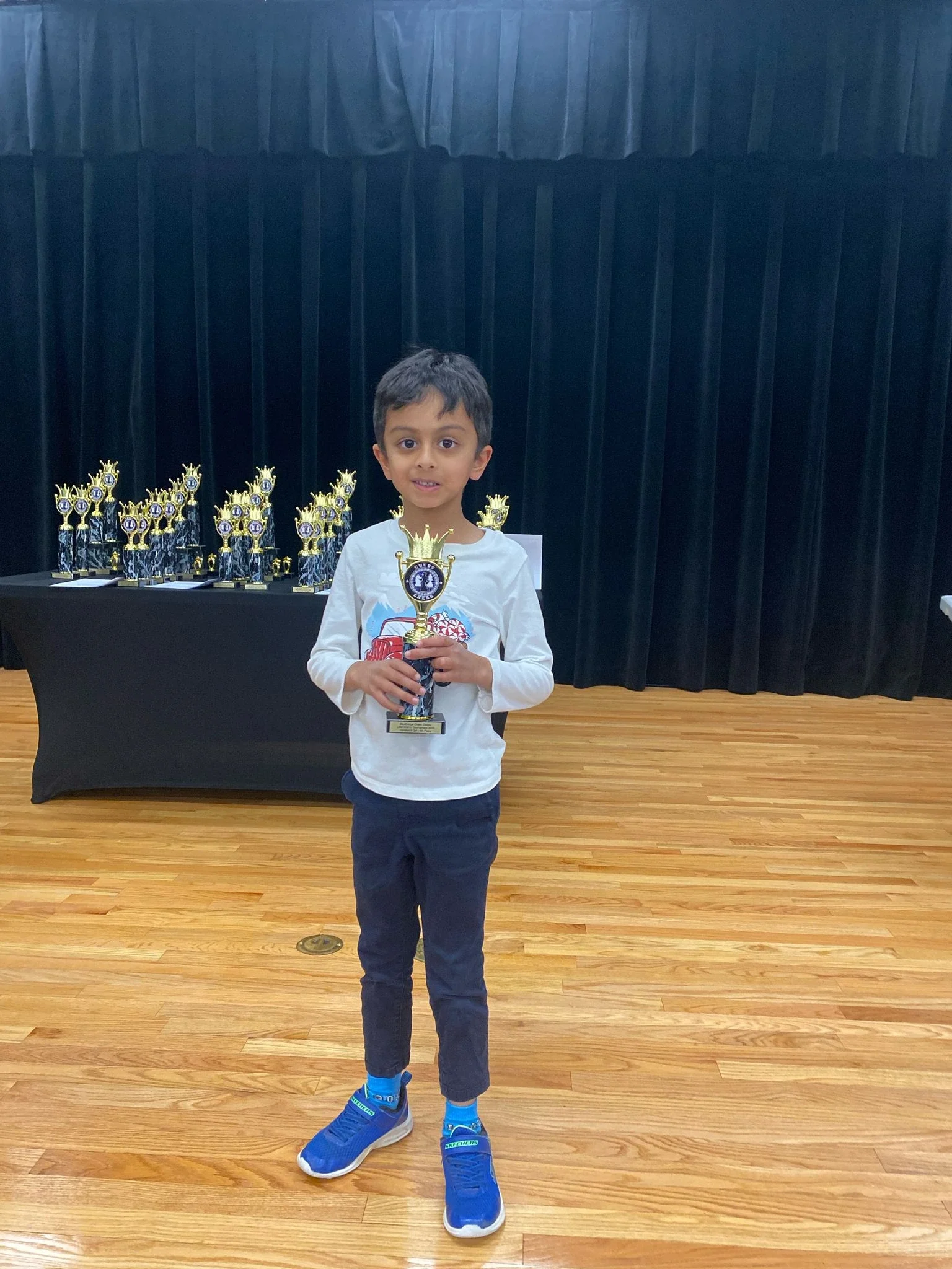 A young boy smiling and holding his chess trophy, with a display table full of similar chess awards in the background