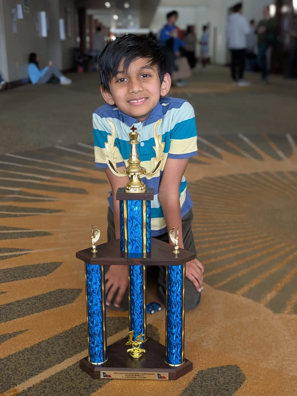 A smiling young boy kneeling behind his first-place blue and gold trophy from the Lone Star Scholastic Chess Championships in Houston.