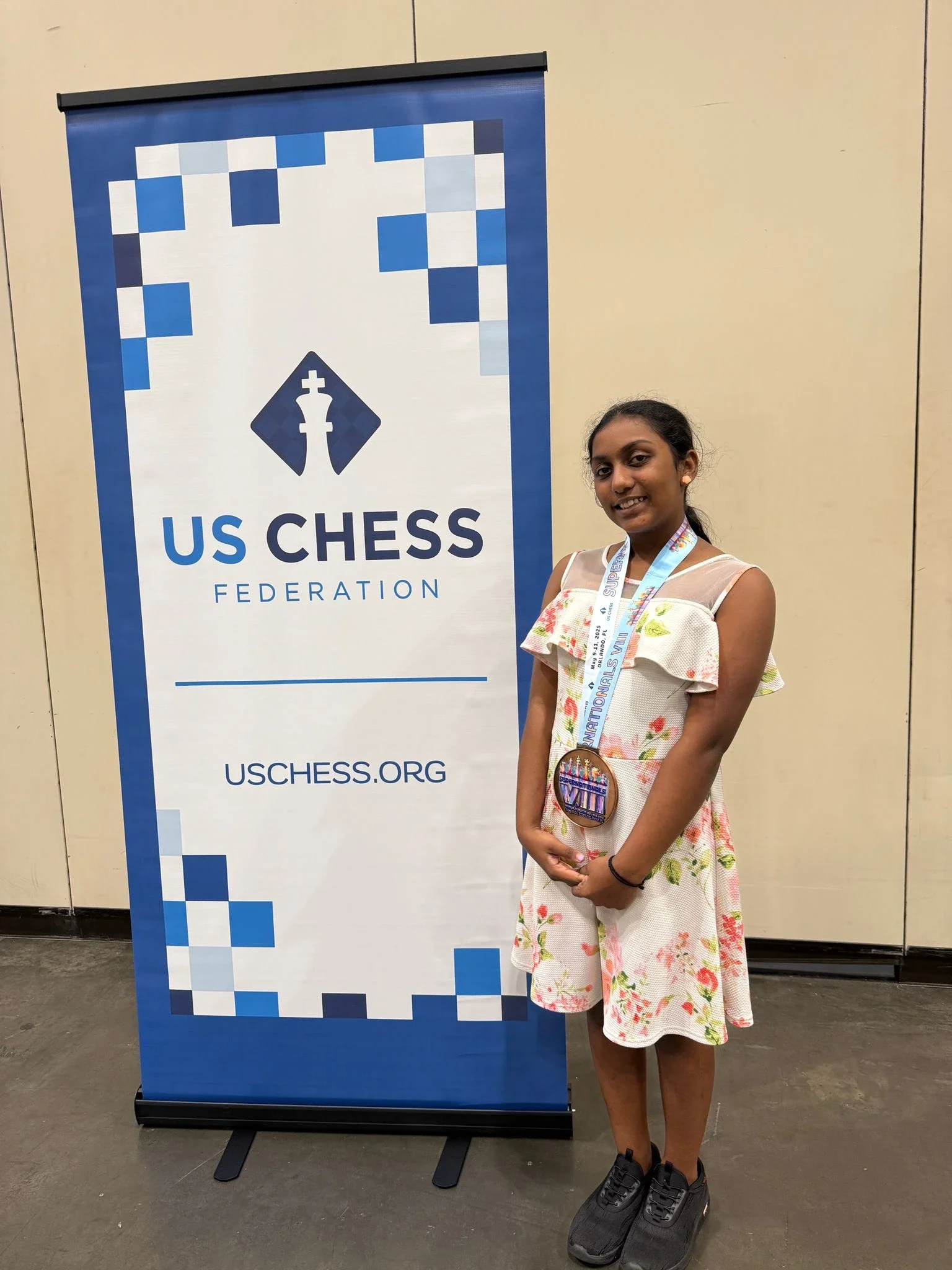 A young girl wearing a large chess tournament medal, posing next to a US Chess Federation banner.