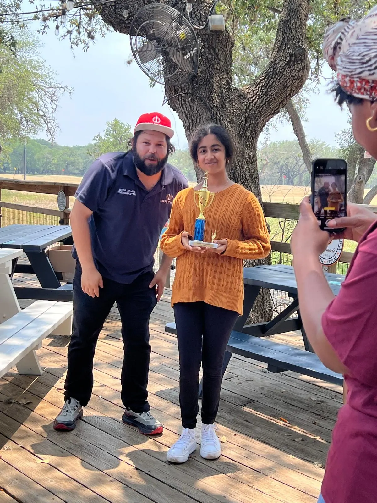 A young girl holding a blue and gold chess trophy while posing for a photo outdoors with a Chess coach.