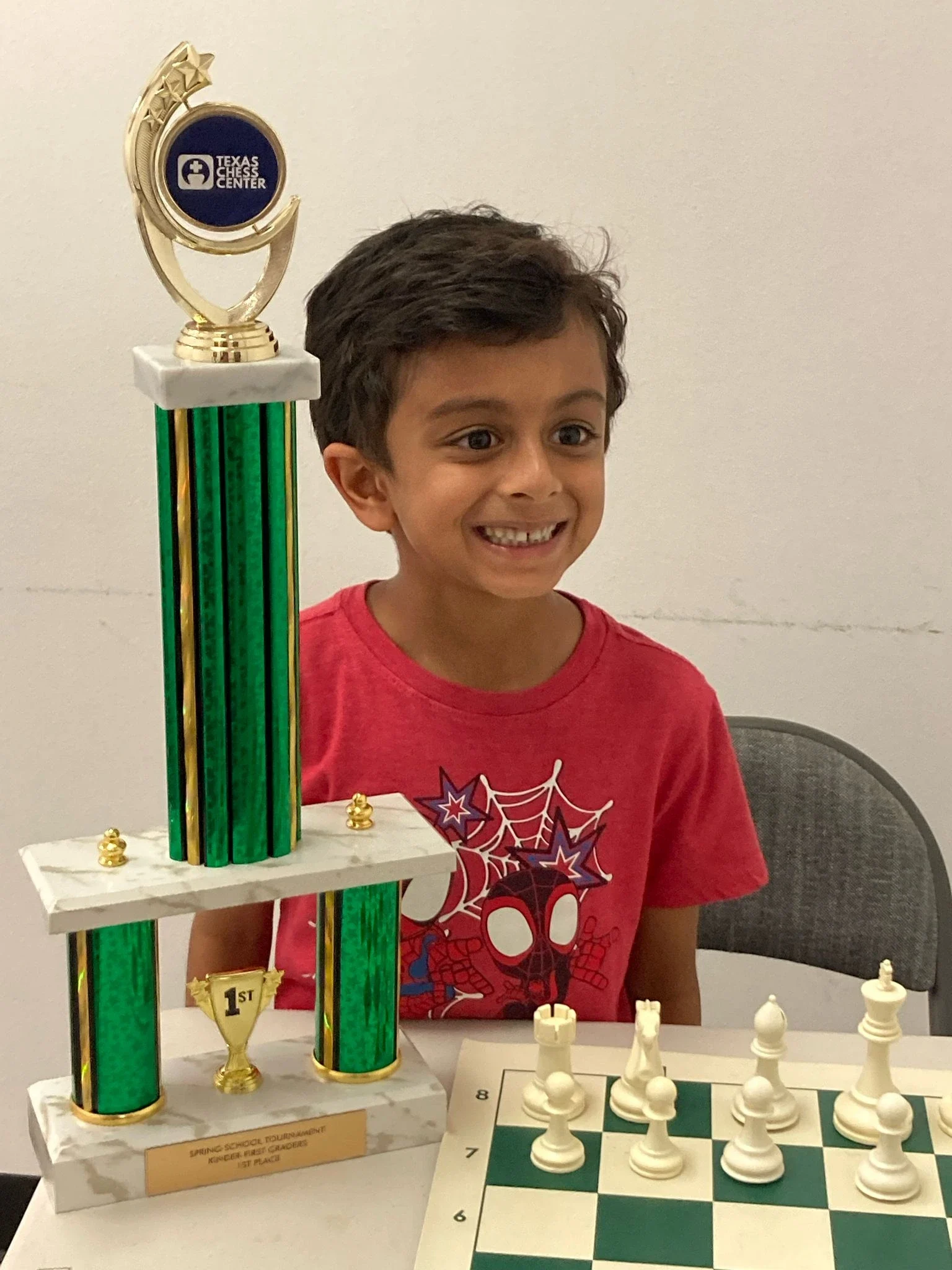 A happy young chess player sitting at a chessboard next to his first-place at texas chess centre spring school tournament which is a tall green trophy